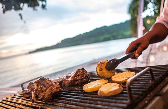 a hand flipping pineapple over on a grill in front of a lake